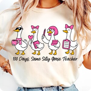 K&ouml;nnte beinhalten: Cremefarbenes T-Shirt mit einem Design von vier Cartoon-G&auml;nsen mit rosa Akzenten. Die G&auml;nse tragen Accessoires wie Sonnenbrillen, Schleifen und B&uuml;cher. Der Text "100 Days, Same Silly Goose Teacher" ist darunter gedruckt.