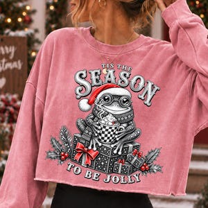 Puede incluir: Una sudadera corta de color rosa empolvado con el gr&aacute;fico de una rana con gorro de Pap&aacute; Noel y gafas, sosteniendo una taza de cacao. El texto 'TIS THE SEASON TO BE JOLLY' rodea a la rana, que est&aacute; sentada sobre cajas de regalo envueltas.