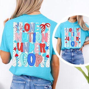 Puede incluir: Una camiseta azul claro con el texto "ONE NATION UNDER GOD" en un dise&ntilde;o patri&oacute;tico colorido. El dise&ntilde;o incluye estrellas rojas, blancas y azules, fuegos artificiales y un patr&oacute;n de cuadros. La camiseta es de estilo casual.