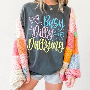 Puede incluir: Sudadera gris oscuro con el texto "Busy Dilly Dallying" en escritura azul, rosa y amarilla, acentuada con gr&aacute;ficos de lazo. Un c&aacute;rdigan de patchwork colorido en rosa, naranja, amarillo y azul se usa sobre la sudadera.
