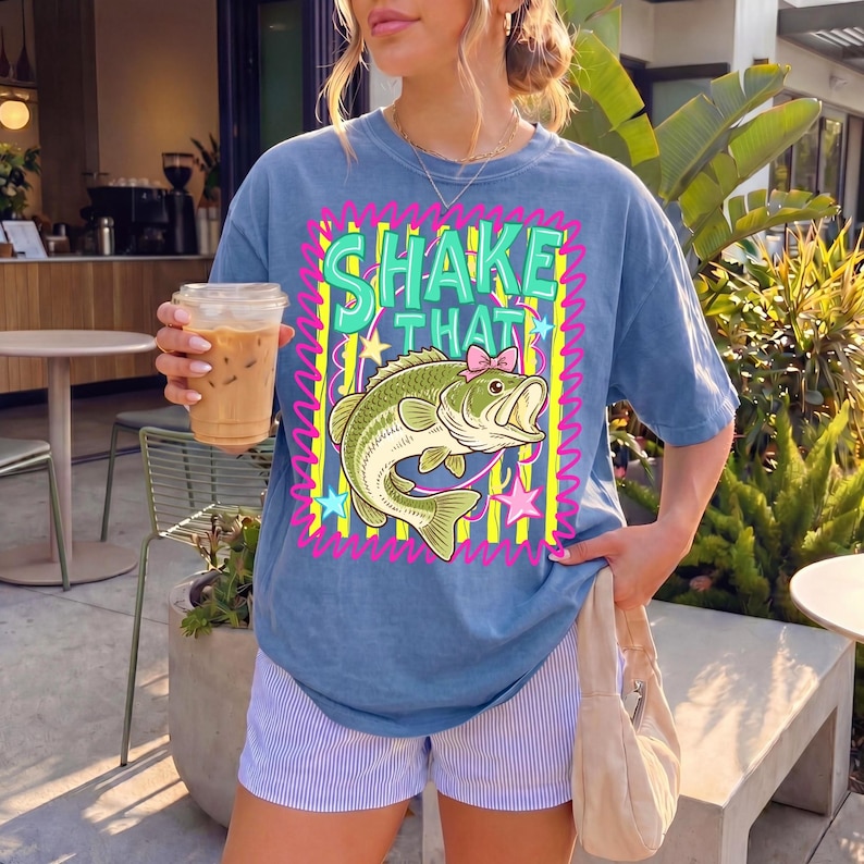 Op de afbeelding: Blauw T-shirt met de woorden "SHAKE THAT" in groene letters boven een cartoonvis met een roze strik. De vis is groen en wit met gele sterren en een roze zigzagrand. De persoon draagt een blauw-witte gestreepte short en houdt een koffiedrankje vast.