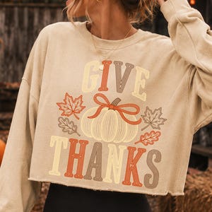 Puede incluir: Una sudadera corta de color beige con mangas largas y un estampado gr&aacute;fico con el texto GIVE THANKS en una fuente retro. El dise&ntilde;o incluye una calabaza central con un lazo rojo y hojas de arce y roble circundantes en tonos rojos y marrones.