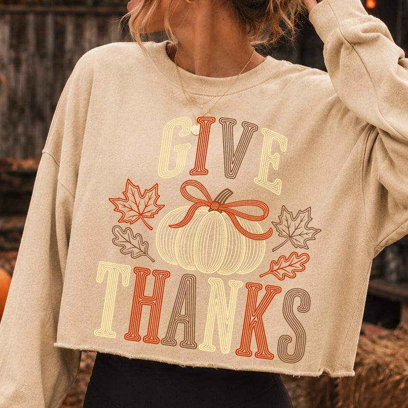 Puede incluir: Una sudadera corta de color beige con mangas largas y un estampado gr&aacute;fico con el texto GIVE THANKS en una fuente retro. El dise&ntilde;o incluye una calabaza central con un lazo rojo y hojas de arce y roble circundantes en tonos rojos y marrones.