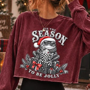 Puede incluir: Una sudadera de manga larga color granate con dobladillo sin rematar, con un gr&aacute;fico de una rana con gorro de Pap&aacute; Noel y gafas, sosteniendo una taza de cacao. El texto TIS THE SEASON TO BE JOLLY est&aacute; rodeado de regalos, acebo y bayas.