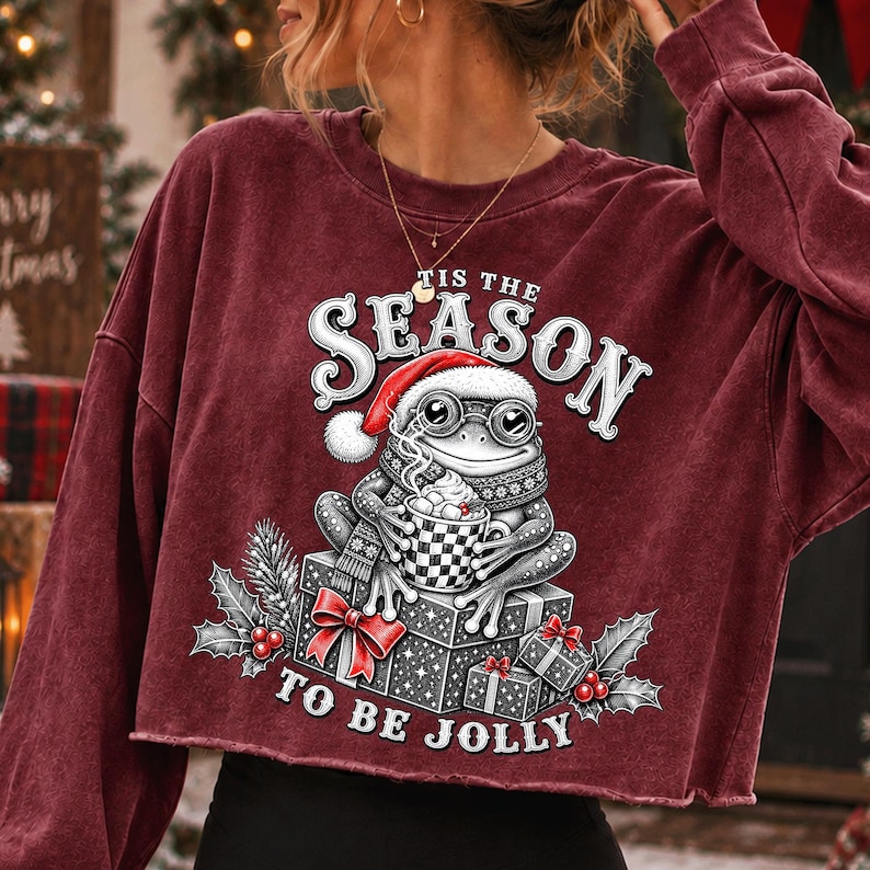 Puede incluir: Una sudadera de manga larga color granate con dobladillo sin rematar, con un gr&aacute;fico de una rana con gorro de Pap&aacute; Noel y gafas, sosteniendo una taza de cacao. El texto TIS THE SEASON TO BE JOLLY est&aacute; rodeado de regalos, acebo y bayas.