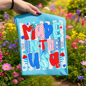 Pode incluir: Uma t-shirt azul-clara com o texto MADE IN THE USA numa fonte estampada multicolorida. As letras est&atilde;o decoradas com bolinhas e padr&otilde;es xadrez vermelhos, brancos e azuis, acentuadas por pequenas ilustra&ccedil;&otilde;es de fogo de artif&iacute;cio e estrelas.