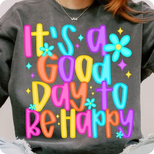 K&ouml;nnte beinhalten: Ein graues Sweatshirt mit dem Text "It's a good day to be happy" in bunten, umrandeten Buchstaben. Der Text ist von kleinen, bunten Blumen und Sternen umgeben.