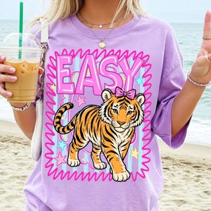 May include: A lilac t-shirt with a cartoon tiger wearing a pink bow, framed by pink wavy lines and the word "EASY". The tiger has orange and black stripes. The background has blue stripes and stars. The t-shirt is a relaxed fit.