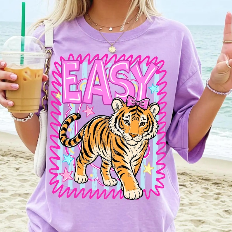 May include: A lilac t-shirt with a cartoon tiger wearing a pink bow, framed by pink wavy lines and the word "EASY". The tiger has orange and black stripes. The background has blue stripes and stars. The t-shirt is a relaxed fit.