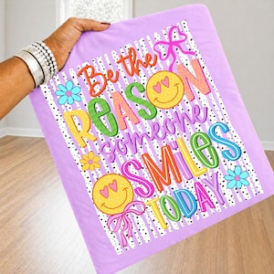 May include: A lavender blanket with the text "Be the REASON someone SMILES TODAY" in colorful letters. The design includes smiley faces, flowers, and bows. The blanket is held up by a person.