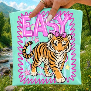 May include: A light teal tote bag with a cartoon tiger in orange and black stripes, a pink bow, and the word "EASY" in pink bubble letters. The design includes pink stars and a pink wavy border.