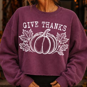 Puede incluir: Una sudadera de color ciruela con cuello redondo y mangas largas, con un dobladillo corto de borde sin rematar. El frente presenta un estampado gr&aacute;fico blanco de una calabaza flanqueada por hojas de arce y roble, con el texto GIVE THANKS.