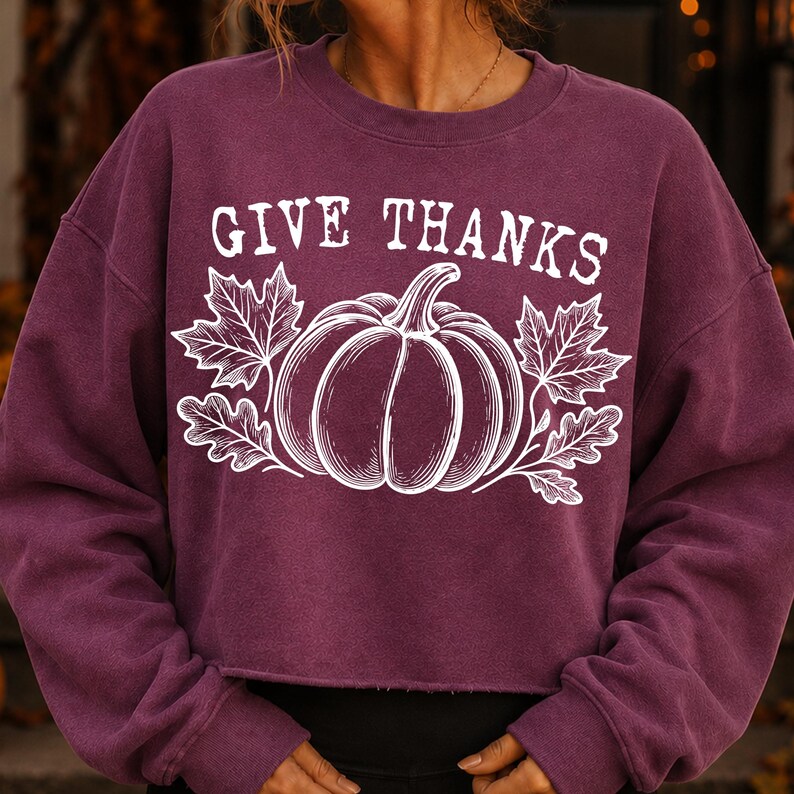 Puede incluir: Una sudadera de color ciruela con cuello redondo y mangas largas, con un dobladillo corto de borde sin rematar. El frente presenta un estampado gr&aacute;fico blanco de una calabaza flanqueada por hojas de arce y roble, con el texto GIVE THANKS.