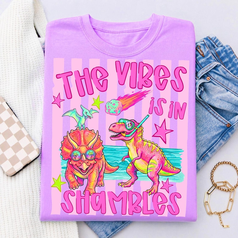 Peut inclure: T-shirt lilas avec un motif de dinosaures de dessins anim&eacute;s portant des lunettes de soleil et un &eacute;quipement de plong&eacute;e. Le texte "THE VIBES IS IN SHAMBLES" est en rose. Le design comprend des &eacute;toiles et une com&egrave;te.