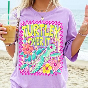 Puede incluir: Camiseta lila claro con las palabras "TURTLEY OVER IT" en amarillo y rosa. La camiseta presenta una tortuga turquesa con flores rosas, amarillas y naranjas. Corte holgado.