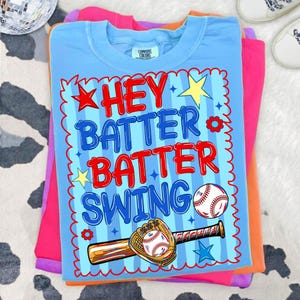 K&ouml;nnte beinhalten: Ein Stapel bunter T-Shirts, mit einem hellblauen Shirt oben. Das hellblaue Shirt tr&auml;gt die Worte "HEY BATTER BATTER SWING" in Rot und Blau, mit Baseball-Grafiken. Die anderen Shirts sind rosa und orange.