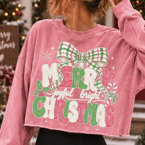 K&ouml;nnte beinhalten: Ein altrosa lang&auml;rmeliges bauchfreies Sweatshirt mit einem gro&szlig;en Aufdruck einer gr&uuml;n-wei&szlig; karierten Schleife und einer silbernen Glocke. Der Schriftzug Merry joyful & bright Christmas ist in Rot, Gr&uuml;n und Wei&szlig; gehalten und mit Stechpalmen verziert.