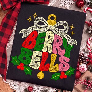 May include: A black t-shirt featuring a graphic of a Christmas ornament shaped like a bell with a bow on top. The text "BERRY BELLS" is written in a stylized, scribbled font in red, green, pink, and yellow. Green holly leaves and red berries are at the bottom.
