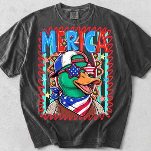 May include: Dark gray t-shirt featuring a cartoon duck wearing star-shaped sunglasses, a baseball cap, and a bandana in red, white, and blue. The word "MERICA" is displayed in a colorful, patriotic design.