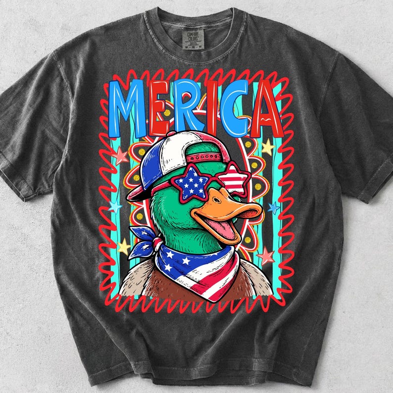 May include: Dark gray t-shirt featuring a cartoon duck wearing star-shaped sunglasses, a baseball cap, and a bandana in red, white, and blue. The word "MERICA" is displayed in a colorful, patriotic design.