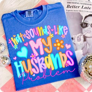 K&ouml;nnte beinhalten: Ein blaues T-Shirt mit dem Schriftzug "that sounds like MY husbands problem" in bunten, verspielten Buchstaben. Das Design umfasst Herzen und Blumen. Das Shirt ist von der Marke Comfort Colors.