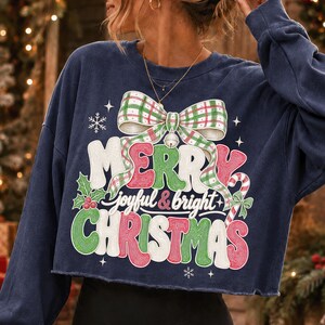 K&ouml;nnte beinhalten: Ein marineblaues, bauchfreies Langarm-Sweatshirt mit einem Grafikdruck aus einer karierten Schleife, einer Zuckerstange, Stechpalmenbeeren und dem Text Merry joyful & bright CHRISTMAS in Wei&szlig;, Rot und Gr&uuml;n mit kleinen Schneeflocken.