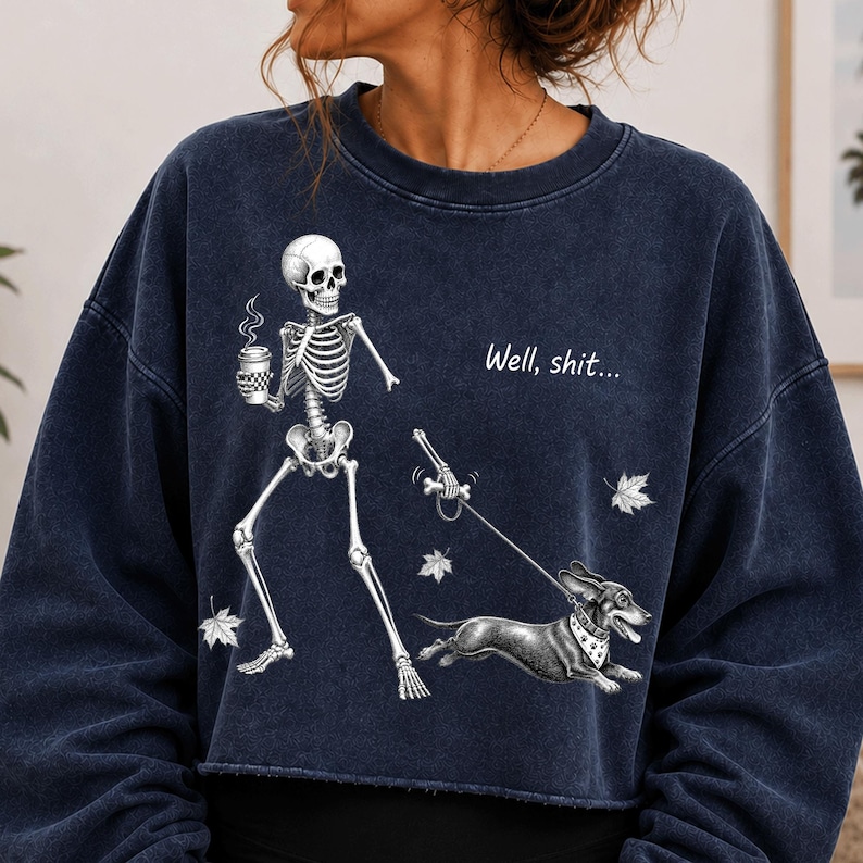 May include: A dark blue mineral-washed crewneck sweatshirt featuring a graphic of a skeleton holding a coffee cup and walking a dachshund on a leash. The text "Well, shit..." is printed above the dog, accompanied by scattered white maple leaves.