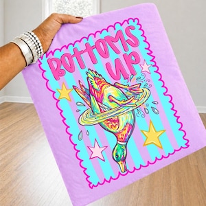 May include: A light purple blanket with a teal border and vertical stripes. The blanket features a colourful illustration of a duck diving into water, with the words "BOTTOMS UP" in pink.