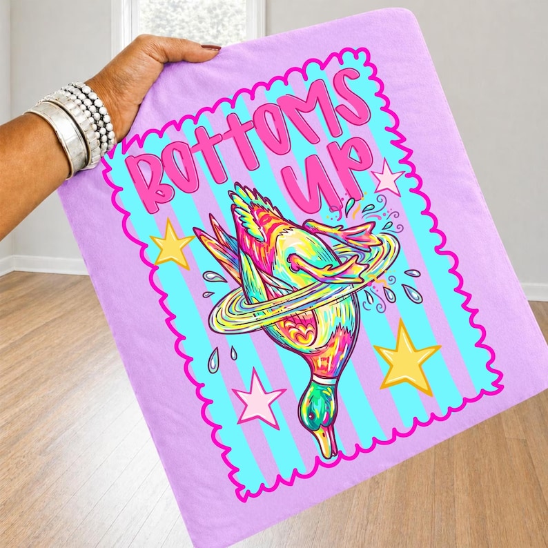 May include: A light purple blanket with a teal border and vertical stripes. The blanket features a colourful illustration of a duck diving into water, with the words "BOTTOMS UP" in pink.