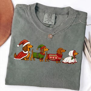 May include: A grey t-shirt featuring a graphic of four dachshunds dressed in holiday attire. One wears a Santa hat and cape, one an elf hat and tunic, one a red patterned jumper, and one a white hooded cloak with holly accents.