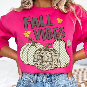Puede incluir: Una sudadera de manga larga en color rosa brillante con el texto FALL VIBES en letras gris oscuro. Debajo del texto hay tres calabazas con un patr&oacute;n de cuadros y camuflaje, acentuadas por peque&ntilde;as ilustraciones de hojas amarillas y grises.