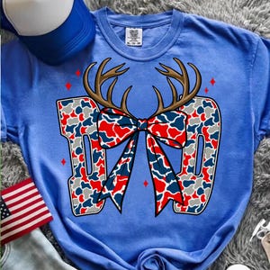 以下が含まれることがあります： 赤、白、青のカモフラージュ柄で「DAD」の文字が入った青いTシャツ。同じ模様のリボンが中央にあり、鹿の角が上にあります。赤いひし形がデザインの周りに散らばっています。