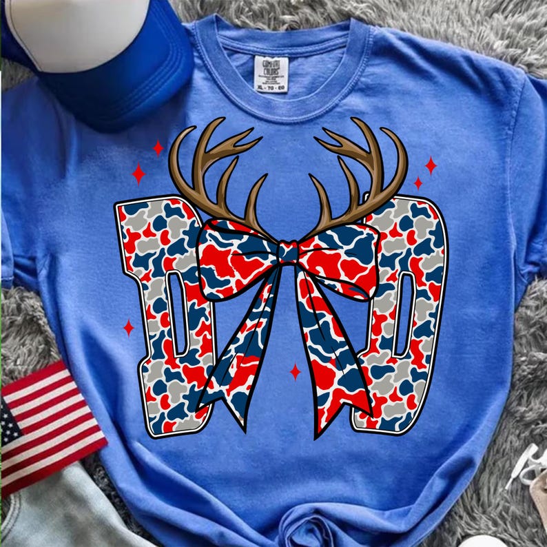 以下が含まれることがあります： 赤、白、青のカモフラージュ柄で「DAD」の文字が入った青いTシャツ。同じ模様のリボンが中央にあり、鹿の角が上にあります。赤いひし形がデザインの周りに散らばっています。