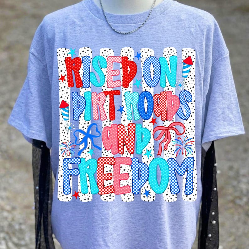 Op de afbeelding: Een lichtgrijs t-shirt met korte mouwen en een grafische print met de tekst "RISED ON DIRT ROADS AND FREEDOM". Het ontwerp bevat rode, witte en blauwe patronen, stippen, strepen, vuurwerkillustraties en strikken op een witte achtergrond.