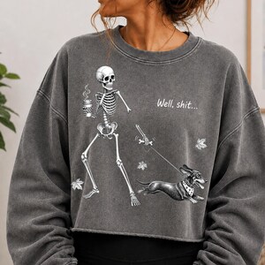 May include: A charcoal gray cropped sweatshirt featuring a graphic of a skeleton holding a coffee cup and walking a dachshund on a leash. The skeleton is holding a bone, and the text "Well, shit..." is printed above the dog, with scattered leaves.