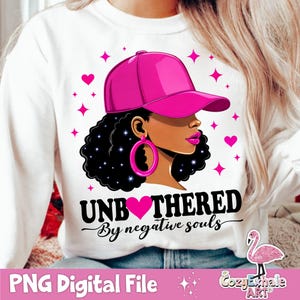 May include: A white sweatshirt with a pink and black graphic design. The graphic features a woman with a pink baseball cap, black hair, and a pink heart. The text "Unbothered by negative souls" is written in black below the graphic.
