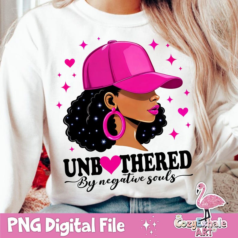 May include: A white sweatshirt with a pink and black graphic design. The graphic features a woman with a pink baseball cap, black hair, and a pink heart. The text "Unbothered by negative souls" is written in black below the graphic.