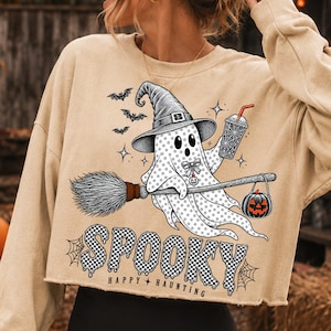 May include: A beige cropped sweatshirt featuring a graphic of a ghost wearing a witch hat, holding a drink and a broomstick with a jack-o'-lantern. The design includes bats, stars, and the text "SPOOKY HAPPY HAUNTING" in a black and white checkered pattern.