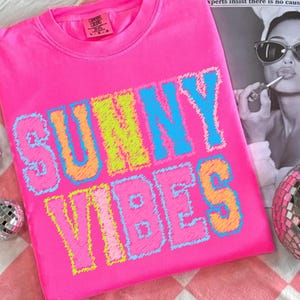 May include: Bright pink crew neck sweatshirt with the words "SUNNY VIBES" in a colorful, stitched font. The letters are outlined in blue, yellow, and orange. The sweatshirt has a Comfort Colors tag.