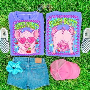 Op de afbeelding: Een platte lay-out met een lila t-shirt met een varkensafbeelding met een hartvormige zonnebril en de tekst "GUESS WHAT?" en "PIGGY BUTT!". Ook inbegrepen zijn een spijkerbroek, een roze truckerpet, een blauwe scrunchie en geruite slip-on sneakers.