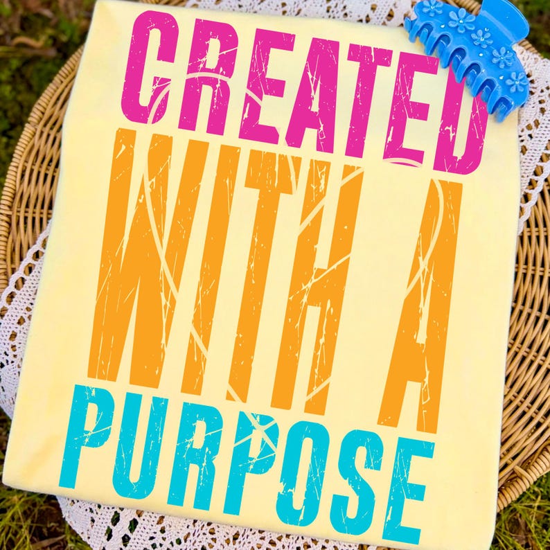 May include: A light yellow t-shirt with the words "CREATED WITH A PURPOSE" in pink, orange, and teal. The shirt is on a wicker basket with a white lace doily. A blue hair clip with flower details rests on the shirt.