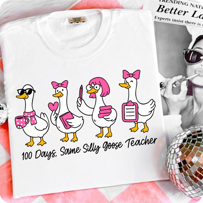 K&ouml;nnte beinhalten: Wei&szlig;es T-Shirt mit einem Cartoon-Design von vier G&auml;nsen mit rosa Accessoires, die jeweils verschiedene Gegenst&auml;nde halten. Der Text "100 Days, Same Silly Goose Teacher" ist darunter gedruckt. Das Shirt liegt auf einer rosa und wei&szlig; gestreiften Oberfl&auml;che.