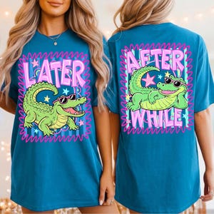 May include: Teal t-shirt featuring a cartoon alligator wearing sunglasses. The front of the shirt says "Later" and the back says "After While". The design is framed in a pink and blue border.