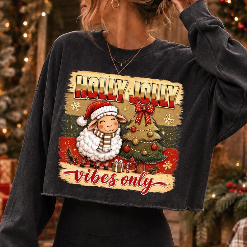 Może przedstawiać: Ciemnoszara bluza z długim rękawem z nadrukiem owcy w czerwonej czapce Mikołaja i szaliku obok udekorowanej choinki. Napis HOLLY JOLLY vibes only wykonany jest czerwoną i złotą czcionką na tle w paski, co tworzy świąteczny motyw graficzny.