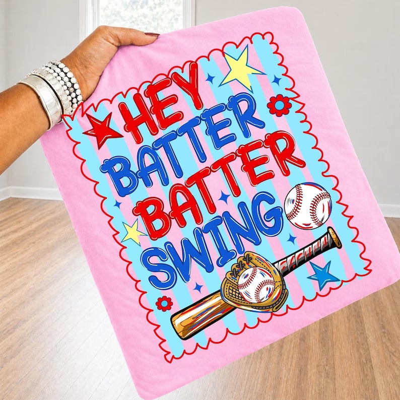 K&ouml;nnte beinhalten: Rosa Baseball-Decke mit den Worten "Hey Batter Batter Swing" in Rot und Blau. Das Design umfasst einen Baseball, einen Schl&auml;ger und einen Handschuh, mit Stern- und Blumenakzenten. Die Decke wird von einer Person gehalten.