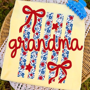 Op de afbeelding: Een lichtgeel t-shirt met het woord "grandma" in rood script. Het shirt heeft verticale strepen met een bloemenpatroon in rood, wit en blauw, samen met rode strikken en sterren. Een blauwe haarclip bevindt zich in de rechterbovenhoek.