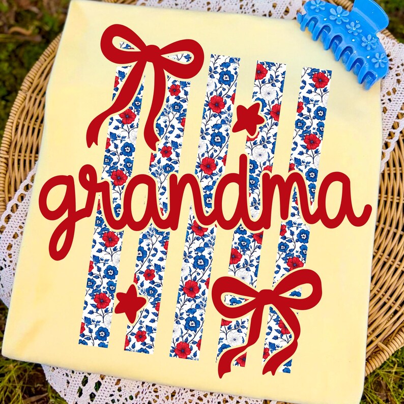 Op de afbeelding: Een lichtgeel t-shirt met het woord "grandma" in rood script. Het shirt heeft verticale strepen met een bloemenpatroon in rood, wit en blauw, samen met rode strikken en sterren. Een blauwe haarclip bevindt zich in de rechterbovenhoek.
