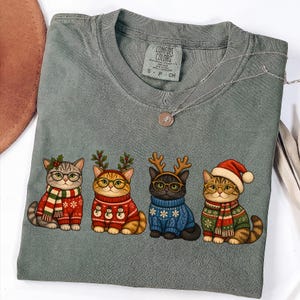 K&ouml;nnte beinhalten: Ein salbeigr&uuml;nes T-Shirt mit Rundhalsausschnitt und einer Grafik von vier Katzen in festlicher Kleidung. Die Katzen tragen Pullover, Schals und weihnachtliche Kopfbedeckungen wie Rentiergeweihe und eine Weihnachtsmannm&uuml;tze sowie runde Brillen.