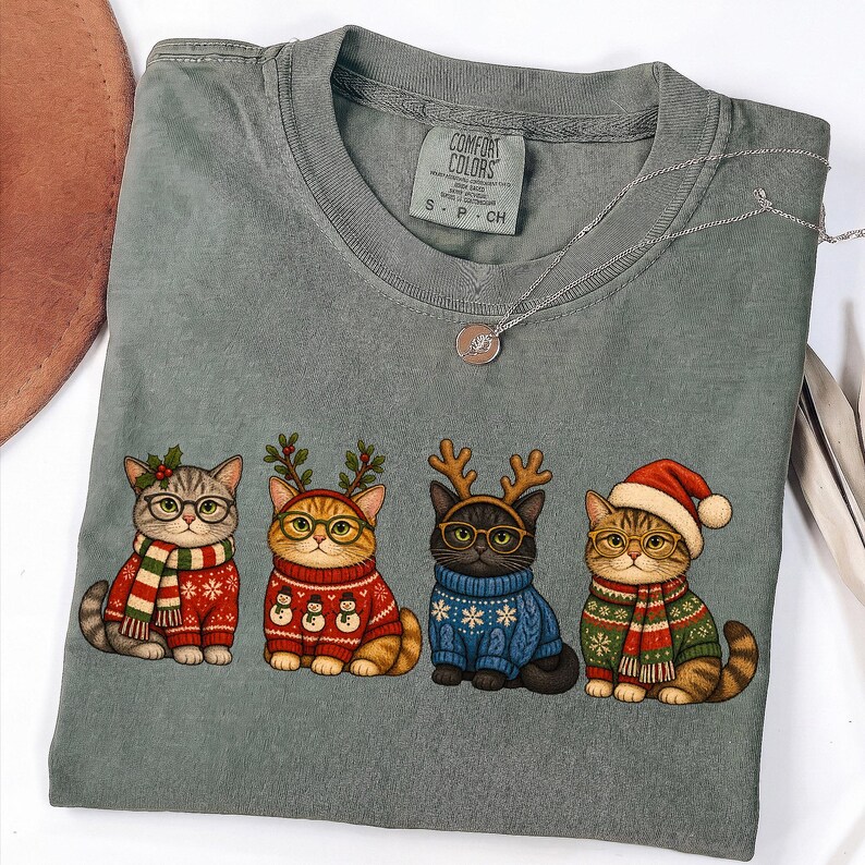K&ouml;nnte beinhalten: Ein salbeigr&uuml;nes T-Shirt mit Rundhalsausschnitt und einer Grafik von vier Katzen in festlicher Kleidung. Die Katzen tragen Pullover, Schals und weihnachtliche Kopfbedeckungen wie Rentiergeweihe und eine Weihnachtsmannm&uuml;tze sowie runde Brillen.