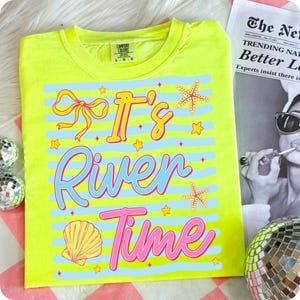May include: A neon yellow t-shirt with the phrase "It's River Time" in a playful font. The design includes a bow, starfish, a seashell, and stars, all set against blue horizontal stripes. The shirt is a Comfort Colors brand.